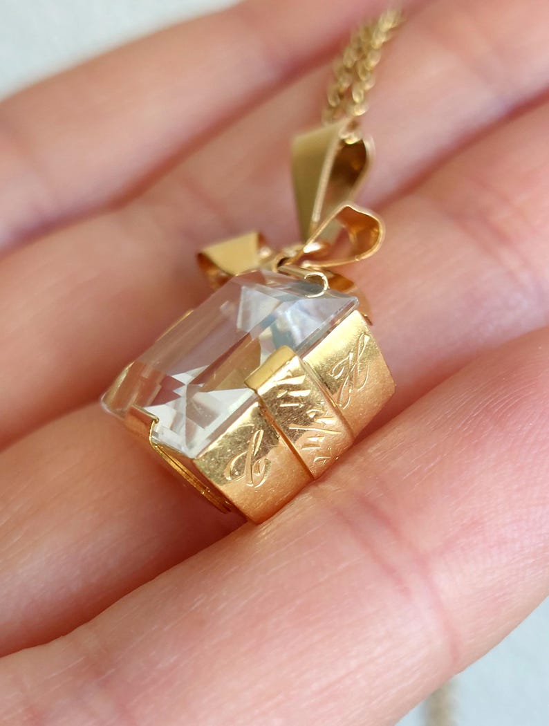 Op de afbeelding: Een goudkleurige hangerketting met een heldere, rechthoekige kristal. De kristal is gezet in een gouden frame met een strikvormige balein. Het gouden frame heeft gegraveerde details. De ketting wordt in een hand gehouden.
