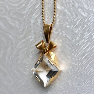 Op de afbeelding: Een goudkleurige ketting met een heldere, facetgeslepen, diamantvormige hanger. De hanger hangt aan een fijne gouden ketting. De hanger is gezet in goud met een strikvormig detail aan de bovenkant. De achtergrond is een witte, getextureerde ondergrond.