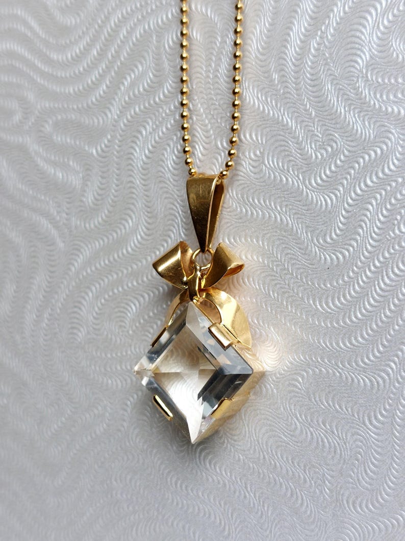 Op de afbeelding: Een goudkleurige ketting met een heldere, facetgeslepen, diamantvormige hanger. De hanger hangt aan een fijne gouden ketting. De hanger is gezet in goud met een strikvormig detail aan de bovenkant. De achtergrond is een witte, getextureerde ondergrond.