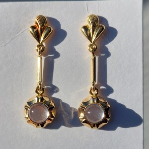 May include: A pair of gold-toned drop earrings featuring a fan-shaped top post connected to a vertical bar and a circular base. Each earring is set with a smooth, translucent, pale pink cabochon stone centered within a scalloped gold metal frame.