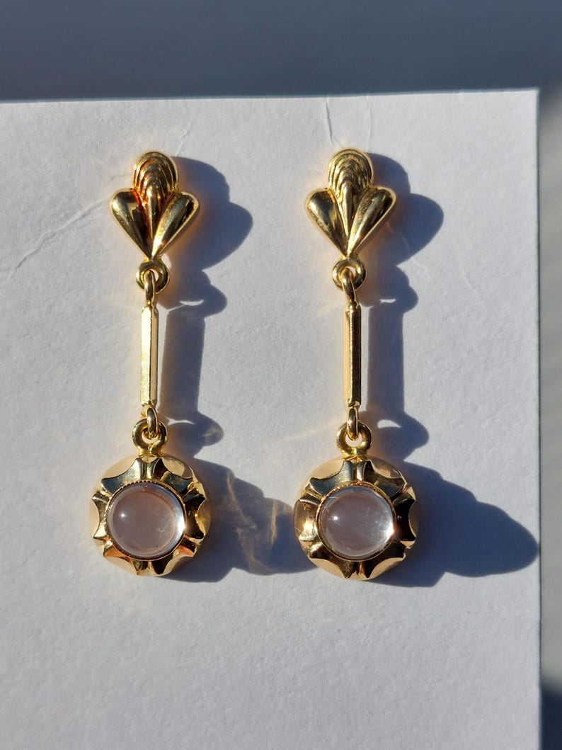 May include: A pair of gold-toned drop earrings featuring a fan-shaped top post connected to a vertical bar and a circular base. Each earring is set with a smooth, translucent, pale pink cabochon stone centered within a scalloped gold metal frame.