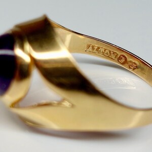 May include: A gold ring with a large, round, deep purple gemstone. The ring has a unique, flowing design with the word "ALTON" stamped on the band. The ring is a close-up shot, highlighting the details of the jewelry.