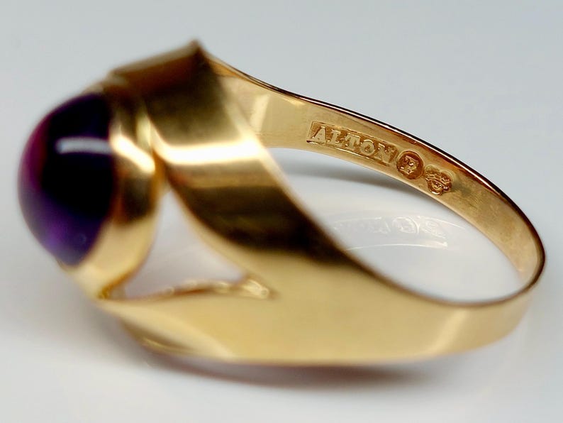 May include: A gold ring with a large, round, deep purple gemstone. The ring has a unique, flowing design with the word "ALTON" stamped on the band. The ring is a close-up shot, highlighting the details of the jewelry.