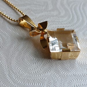 Op de afbeelding: Een gouden hangerketting met een grote, heldere, rechthoekige edelsteen. De hanger is gezet in een gouden frame met een decoratief strikdetail. De ketting is een delicate gouden kogelketting.
