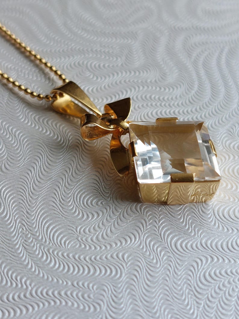 Op de afbeelding: Een gouden hangerketting met een grote, heldere, rechthoekige edelsteen. De hanger is gezet in een gouden frame met een decoratief strikdetail. De ketting is een delicate gouden kogelketting.