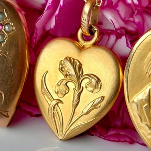 May include: A gold heart-shaped locket pendant featuring a raised relief of an iris flower on the front. The locket is suspended from a gold chain and includes a small bail stamped with the text FIX at the top of the heart-shaped metal piece.
