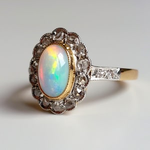 May include: An oval opal ring with a halo of small diamonds. The opal has a rainbow iridescence. The ring's setting is gold and silver, with a gold band. The ring is set against a white background.
