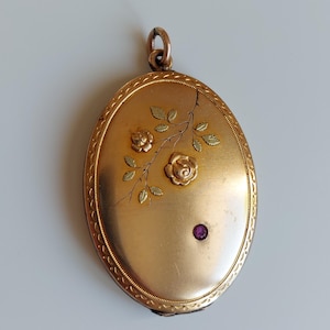 May include: Oval gold locket with floral detailing and a small purple gemstone. The locket has a decorative edge and a loop for a chain. The floral design includes two roses and leaves.