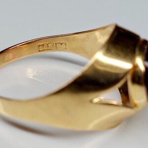 May include: A gold ring with a large, oval, dark purple gemstone. The ring has a thick, polished band and a simple, elegant design. The ring is likely made of gold and features a smooth, reflective surface.