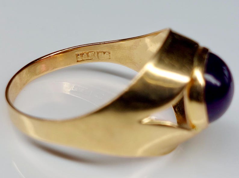 May include: A gold ring with a large, oval, dark purple gemstone. The ring has a thick, polished band and a simple, elegant design. The ring is likely made of gold and features a smooth, reflective surface.