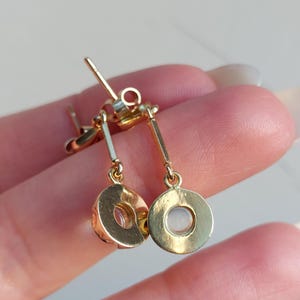 May include: A pair of gold-toned drop earrings featuring a thin vertical bar attached to a circular pendant. Each circular pendant contains a small, translucent, round stone set in the center. The earrings include standard post-style backings.