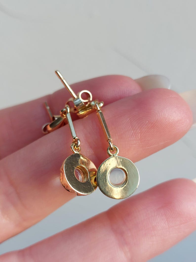 May include: A pair of gold-toned drop earrings featuring a thin vertical bar attached to a circular pendant. Each circular pendant contains a small, translucent, round stone set in the center. The earrings include standard post-style backings.