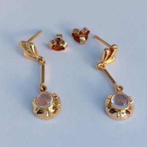 May include: A pair of gold-toned drop earrings featuring a post back and a dangling circular charm. Each charm contains a smooth, translucent, round cabochon stone set within a scalloped metal frame, connected by a thin vertical bar.