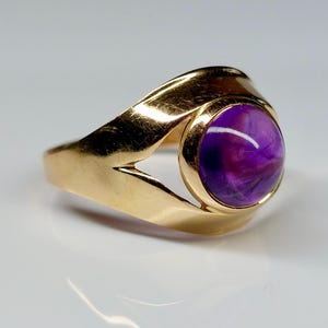 May include: A gold ring featuring a large, round, purple gemstone. The ring has a smooth, polished gold band with a unique, open design on the sides. The gemstone displays a mix of purple hues.