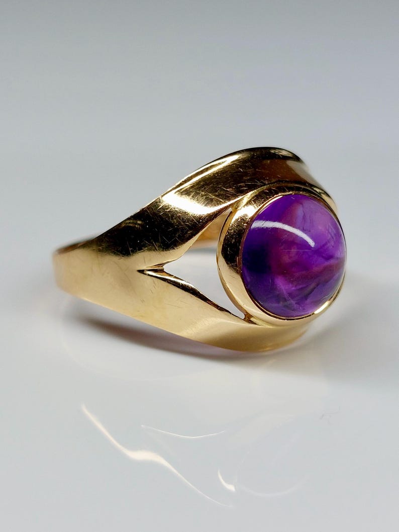May include: A gold ring featuring a large, round, purple gemstone. The ring has a smooth, polished gold band with a unique, open design on the sides. The gemstone displays a mix of purple hues.