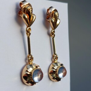 May include: A pair of gold drop earrings featuring a polished metal top section with a vertical bar connecting to a circular base. Each base holds a single, smooth, translucent, pale blue cabochon stone set within a scalloped gold frame.