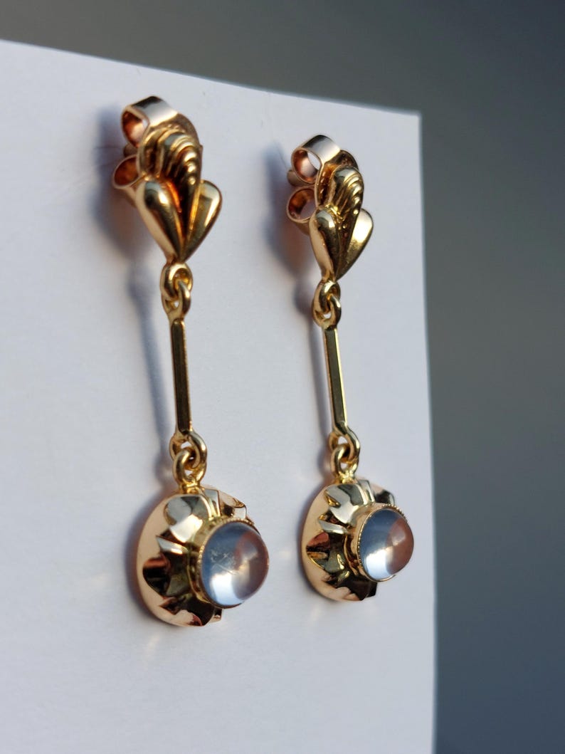 May include: A pair of gold drop earrings featuring a polished metal top section with a vertical bar connecting to a circular base. Each base holds a single, smooth, translucent, pale blue cabochon stone set within a scalloped gold frame.