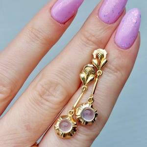 May include: A pair of gold drop earrings featuring a fan-shaped top element connected by a thin bar to a circular base holding a small, translucent, pale pink cabochon stone. The earrings have a polished finish and a classic, structured design.