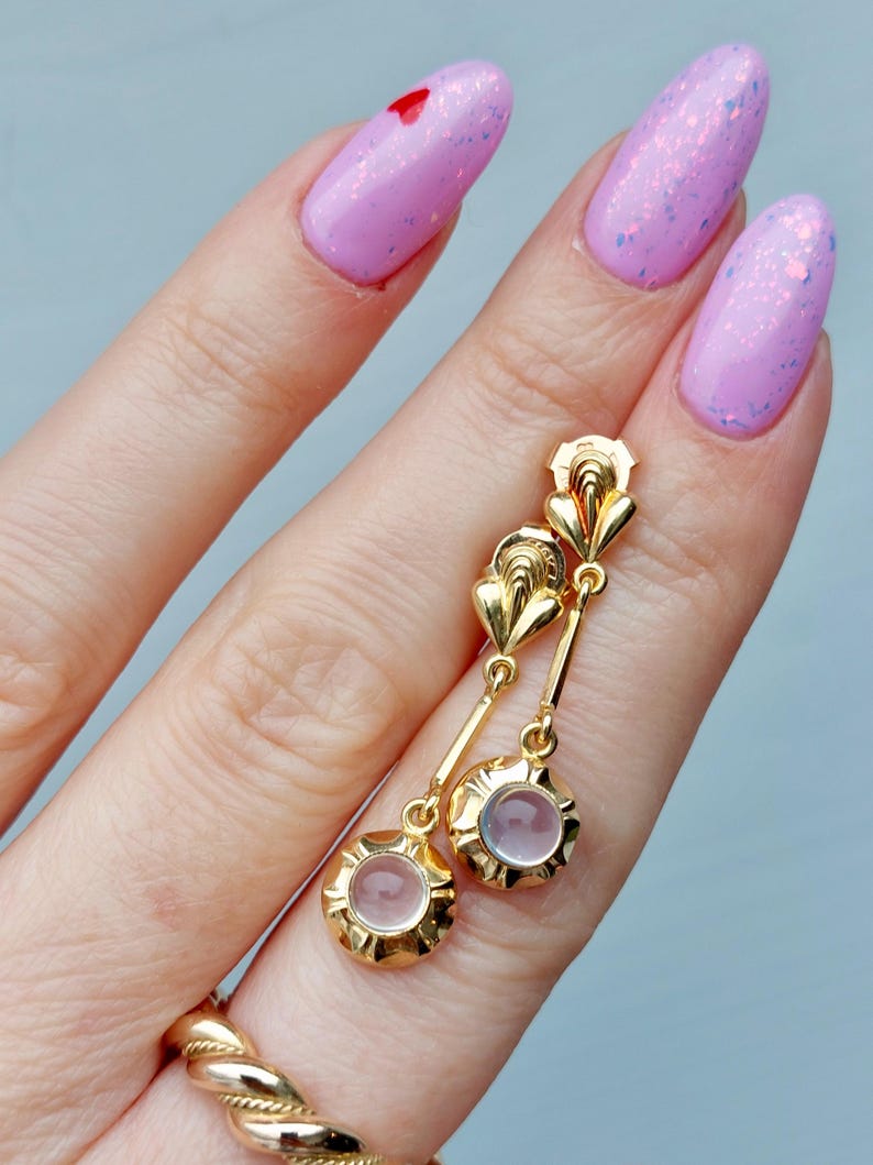 May include: A pair of gold drop earrings featuring a fan-shaped top element connected by a thin bar to a circular base holding a small, translucent, pale pink cabochon stone. The earrings have a polished finish and a classic, structured design.