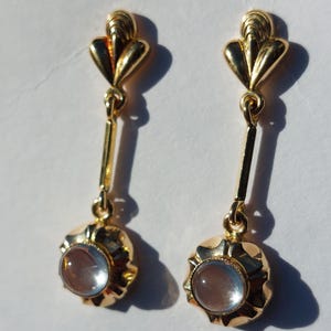 May include: A pair of gold-toned drop earrings featuring a fan-shaped top post connected by a thin vertical bar to a circular base. Each base holds a single, smooth, translucent, light-colored cabochon stone set in a polished metal frame.