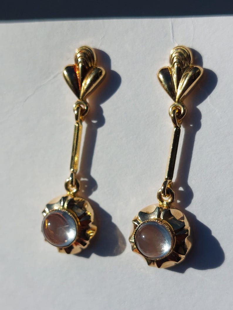 May include: A pair of gold-toned drop earrings featuring a fan-shaped top post connected by a thin vertical bar to a circular base. Each base holds a single, smooth, translucent, light-colored cabochon stone set in a polished metal frame.
