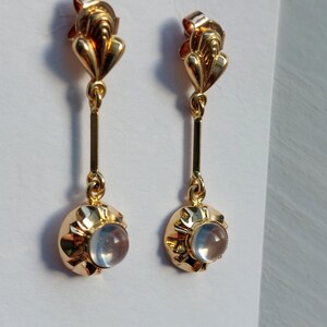 May include: A pair of gold drop earrings featuring a polished gold post with a fan-like top, a vertical gold bar connector, and a circular gold setting holding a smooth, translucent, pale blue cabochon stone at the base of each earring.