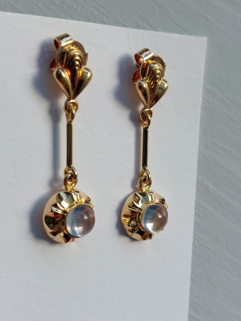 May include: A pair of gold drop earrings featuring a polished gold post with a fan-like top, a vertical gold bar connector, and a circular gold setting holding a smooth, translucent, pale blue cabochon stone at the base of each earring.