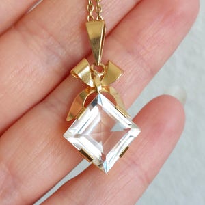 Op de afbeelding: Een goudkleurige ketting met een heldere, vierkant geslepen hanger. De hanger hangt aan een fijne ketting en is versierd met een strikvormig detail. De facetten van de hanger vangen het licht op en cre&euml;ren een sprankelend effect.