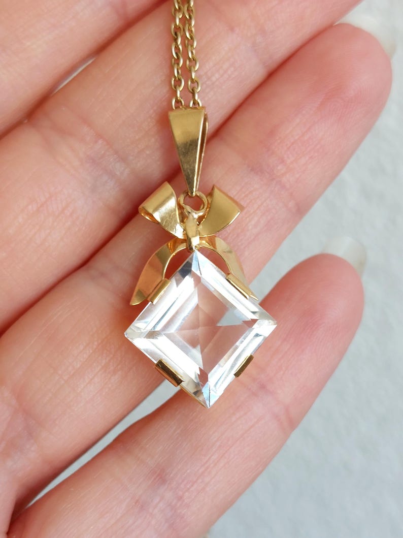 Op de afbeelding: Een goudkleurige ketting met een heldere, vierkant geslepen hanger. De hanger hangt aan een fijne ketting en is versierd met een strikvormig detail. De facetten van de hanger vangen het licht op en cre&euml;ren een sprankelend effect.