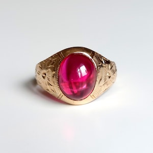 Può includere: Un anello d'oro con una grande gemma ovale rosso rubino. La fascia dell'anello presenta un motivo floreale inciso. La gemma è incastonata in una lunetta d'oro, creando un look classico ed elegante. L'anello è su sfondo bianco.