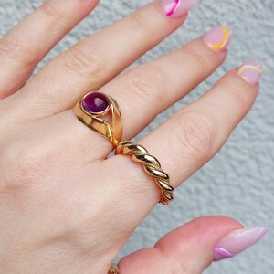 May include: A hand displays three gold rings. One ring features a large, round, purple gemstone. Another is a twisted rope design. The third ring has an engraved floral pattern. The nails are painted with colorful abstract designs.