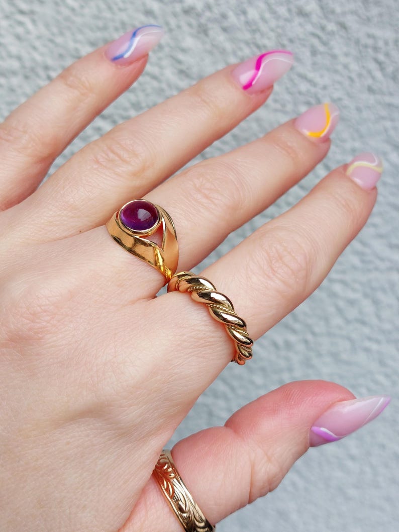 May include: A hand displays three gold rings. One ring features a large, round, purple gemstone. Another is a twisted rope design. The third ring has an engraved floral pattern. The nails are painted with colorful abstract designs.