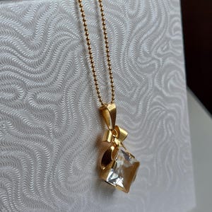 Op de afbeelding: Een gouden ketting met een hanger. De hanger heeft een heldere, vierkant geslepen kristal en een strik ontwerp. De ketting bestaat uit kleine, ronde kralen. De hanger hangt tegen een witte achtergrond.