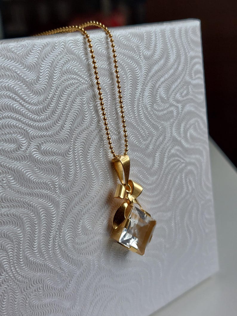 Op de afbeelding: Een gouden ketting met een hanger. De hanger heeft een heldere, vierkant geslepen kristal en een strik ontwerp. De ketting bestaat uit kleine, ronde kralen. De hanger hangt tegen een witte achtergrond.
