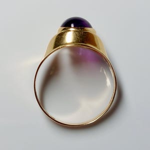 May include: A gold ring with a large, round, deep purple gemstone. The ring's band is a simple, polished gold, and the gemstone is set in a gold bezel. The ring is shown against a white background.