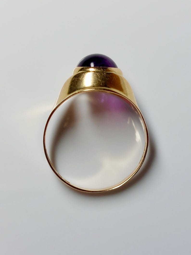 May include: A gold ring with a large, round, deep purple gemstone. The ring's band is a simple, polished gold, and the gemstone is set in a gold bezel. The ring is shown against a white background.