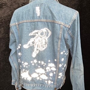 Puede incluir: Chaqueta vaquera azul claro con un gráfico de astronauta blanco en la espalda. La chaqueta presenta un diseño de estrellas, nubes y una luna creciente. El texto en la parte superior de la chaqueta dice "Born Star Sky".