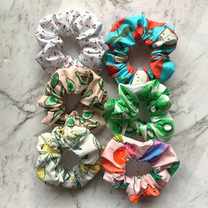 May include: Six colourful scrunchies with different fruit patterns. The scrunchies are made of fabric and have a gathered look. The patterns include cherries, strawberries, avocados, kiwi, and lemons.