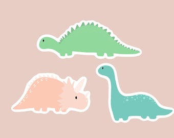 May include: Three colorful dinosaur stickers on a light pink background. The stickers include a green stegosaurus, a peach triceratops, and a teal brontosaurus. Each dinosaur has a white border.
