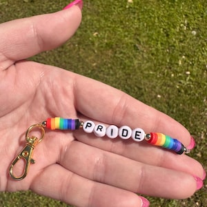 May include: A keychain with the word "PRIDE" spelled out in white letter beads. The keychain features rainbow-colored beads and a gold-colored clasp. The background is a blurred view of green grass.