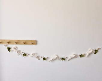 Puede incluir: Una guirnalda de conejos de fieltro blanco con hojas de fieltro verde y cuentas de madera. La guirnalda está enhebrada en una cuerda blanca y está colgada en una pared blanca.
