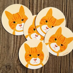 May include: Five round orange and white cat coasters with a cartoon cat face on each. The cat has orange fur, white whiskers, and a white chest. The background of the coasters is a light yellow.