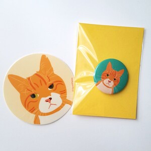 May include: A set of two orange tabby cat illustrations. One is a sticker with a cream background and the other is a button with a teal background. Both feature the same grumpy cat with green eyes.