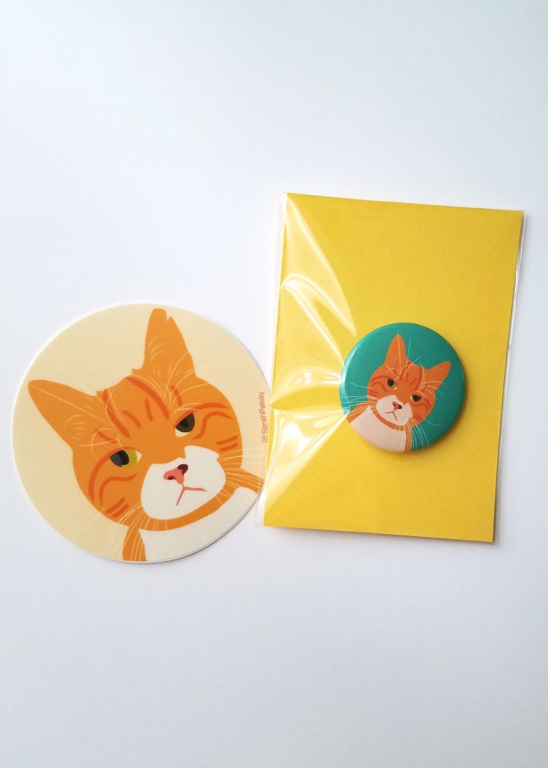 May include: A set of two orange tabby cat illustrations. One is a sticker with a cream background and the other is a button with a teal background. Both feature the same grumpy cat with green eyes.