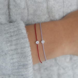 May include: Two delicate bracelets with a small, clear gemstone charm on each. One bracelet is red and the other is light blue.
