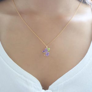 May include: A gold-colored necklace with a pendant featuring purple flower blossoms and green leaves. The necklace is worn against a white top. The pendant is approximately 0.75 inches in length.