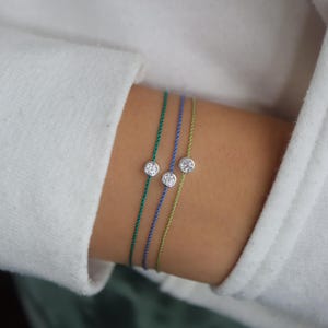 May include: Three fine bracelets, each featuring a single diamond on green, blue, and light green cords. The bracelets are displayed on a wrist, with a white sleeve in the background. The diamonds are set in silver.