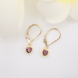 May include: A pair of gold heart-shaped earrings with red gemstone accents. The earrings have a lever back closure.