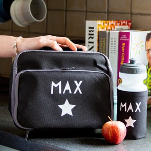 May include: Black lunch bag and water bottle set, both with the name "MAX" and a white star. The lunch bag has a gray zipper and trim. A red apple sits next to the water bottle. The set is designed for carrying food and drinks.