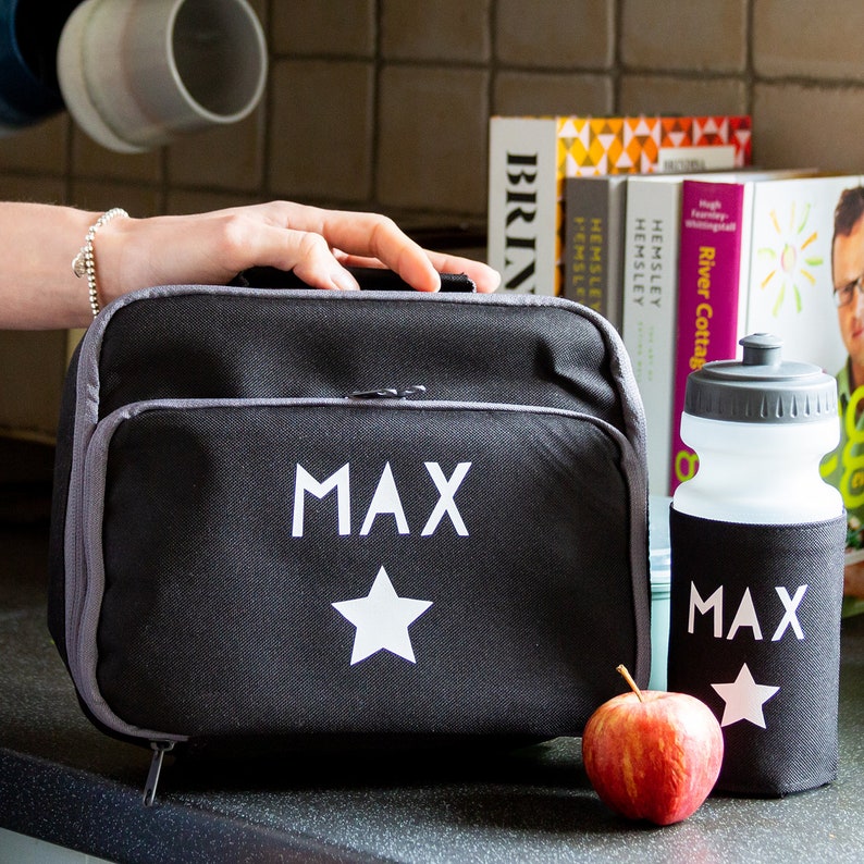 May include: Black lunch bag and water bottle set, both with the name "MAX" and a white star. The lunch bag has a gray zipper and trim. A red apple sits next to the water bottle. The set is designed for carrying food and drinks.